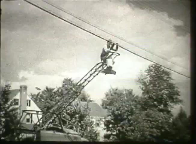 This 16mm still from the Bell System film 'Story Without End' depicts a technician atop a tall, metallic ladder, working on telephone wires. The scene showcases the mid-20th century landscape, featuring lush trees and residential buildings in the background. The image highlights the infrastructure of telecommunications during the late 1940s, emphasizing the hands-on work involved in maintaining the network.