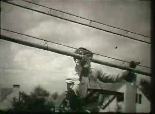 The still from 'Story Without End' features a telephone lineman working on a utility pole, showcasing the infrastructure of the Bell System during the late 1940s. The lineman is wearing safety gear, indicative of the era's emphasis on worker safety, and is engaged in the installation or maintenance of telephone lines. The background includes residential homes, providing a glimpse into the community context that relied on such communication infrastructure. This image underscores the importance of telephone networks in everyday life during that time.
