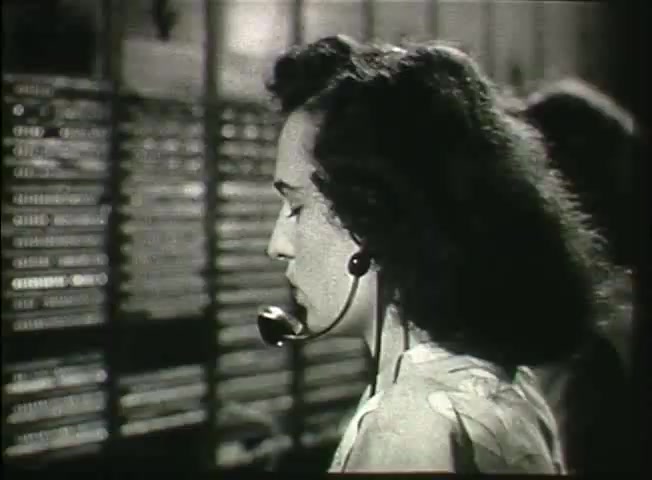 The still from 'Story Without End' depicts a telephone operator in the late 1940s, wearing a headset and focused on her work. She is positioned in front of a large switchboard, which is filled with numerous connections. The image captures the essence of telecommunications during this era, highlighting the human element in connecting calls and managing the network. The setting suggests a busy environment, reflecting the operations of the Bell System at the time.