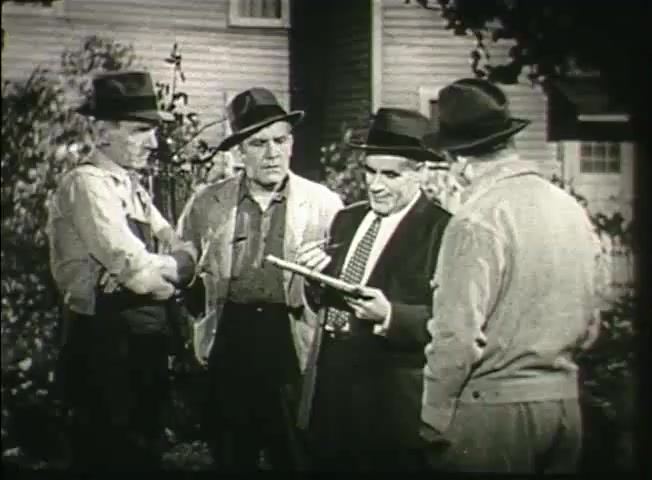 The still from 'Story Without End' features four men engaged in a conversation outdoors, likely discussing technical details related to telecommunications. They are dressed in casual work attire and hats, indicating a mid-20th-century setting. The foreground consists of two men leaning in, focused on a notebook or document held by a third man. The fourth man observes, portraying an atmosphere of collaboration and discussion, typical of Bell System's focus on community and service during the late 1940s. The backdrop suggests a residential area, emphasizing the connection between the network and everyday life.