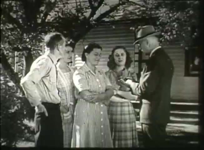 The still from 'Story Without End,' a late 1940s Bell System film, shows a group of four individuals—three women and one man—standing outdoors, engaging with another man in a suit and hat. The setting appears to be a residential area, with a house and trees in the background. The characters’ attire reflects the style of the era, suggesting a casual yet pivotal moment of interaction, possibly discussing telephone services or community connections, which aligns with the film's focus on the telecommunications network of that time.