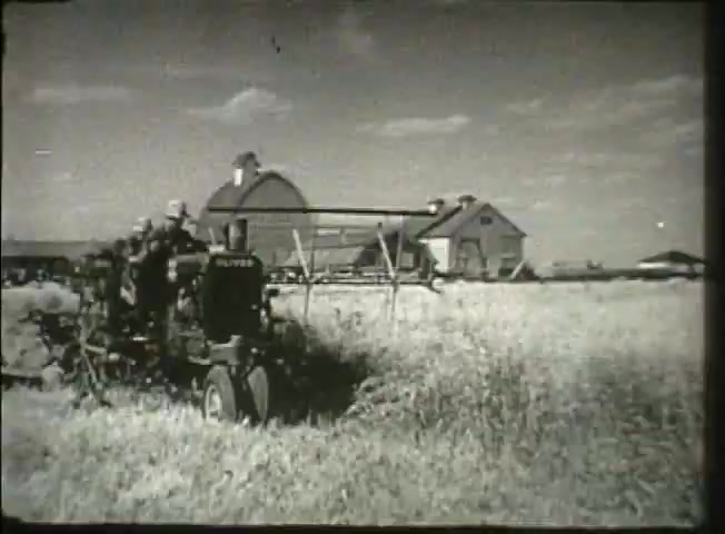 This still from 'Story Without End' features a grain field scene with a farmer operating a tractor and harvesting equipment. In the background, there are two barns under a clear sky, reflecting a rural setting typical of the late 1940s. The image captures the agricultural backdrop that likely emphasizes the theme of communication and modernity in rural America, showcasing how technology was interwoven with everyday farming life during that era.