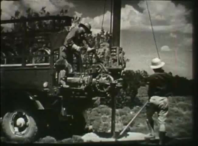 The still from 'Story Without End' depicts two workers engaged in field operations related to the Bell System. One worker is on a truck-like vehicle, possibly operating machinery, while the other stands beside a pole, using a tool to assist with the setup. The background features open terrain under a partly cloudy sky, emphasizing a rural or remote setting. This scene highlights the hands-on aspects of telecommunications infrastructure during the late 1940s, reflecting the era's advancements in network technology.