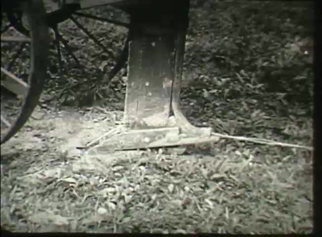 The still shows a close-up of a wooden post or support structure, likely associated with telecommunications infrastructure. It rests on the ground, surrounded by grass, and features a wire extending from it. This image reflects the early telecommunications setup from the late 1940s, emphasizing the foundational elements of the Bell System network.