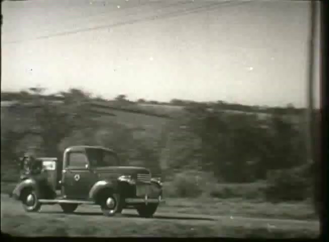 The still depicts a vintage pickup truck driving along a rural road, likely from the late 1940s. The truck, marked with a Bell System logo, suggests its association with telecommunications services. Surrounding greenery hints at a pastoral landscape, highlighting the connection between technology and rural life during that era. The image captures a moment of movement, indicative of service and connectivity in the telecommunications network.