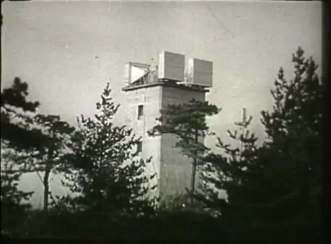 The still from 'Story Without End' features a communication tower surrounded by trees, set against a cloudy sky. The tower has large, square structures on its roof, likely used for transmitting signals. This imagery reflects the technological landscape of the late 1940s, highlighting the infrastructure of the Bell System and its role in communications during that era.
