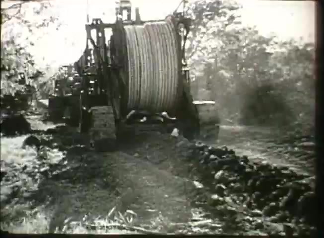 The still depicts heavy machinery, likely a cable reel or plow, used for laying telecommunications lines. The setting suggests an installation phase, possibly in a rural area, showcasing the infrastructure development by Bell System in the late 1940s. Dust and debris indicate active work, emphasizing the technological advancements and efforts in expanding the network during that period.