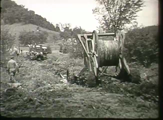 The still from 'Story Without End' features a rural scene with a large cable reel and small trucks, indicative of telecommunications infrastructure work. Workers are seen in the background, illustrating the manual efforts involved in laying down communication lines. The setting reflects the landscape of the late 1940s, showcasing the beginnings of network expansion and the technology of that era.
