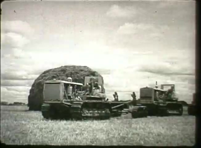 The still from 'Story Without End' features two heavy machinery vehicles, likely tractors, engaged in agricultural work, with a large haystack or similar structure in the background. The setting appears to be a rural landscape under a cloudy sky, suggesting an industrious environment typical of the late 1940s. The focus on machinery highlights advancements in technology during this period, indirectly reflecting the efficiency of the Bell System as a network supporting modern infrastructure.