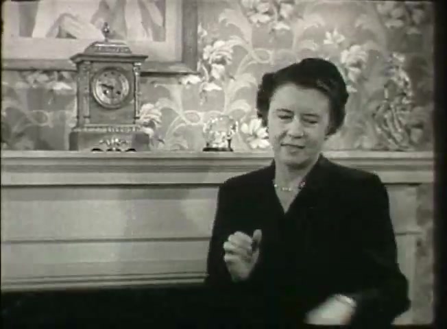 The still captures a woman in a formal black outfit, seated in front of a mantelpiece adorned with a clock and small decorative items. The setting has a vintage aesthetic, featuring patterned wallpaper. The woman appears engaged in conversation, possibly conveying an emotional or thoughtful response. This scene likely relates to themes in the Bell System film 'Story Without End,' which discusses telecommunications and personal connections in the late 1940s.