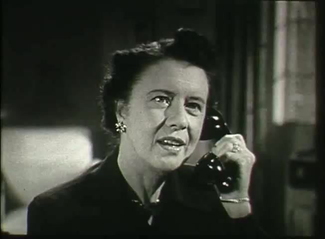 The still features a woman in a late 1940s setting, engaged in a conversation on the phone. She appears focused and thoughtful, suggesting a serious discussion. The black and white aesthetic reflects the era, highlighting the significance of telecommunications in everyday life. The image ties into the themes of the Bell System film 'Story Without End,' which explores the network's role and operations during that time, alongside showcasing central office footage.