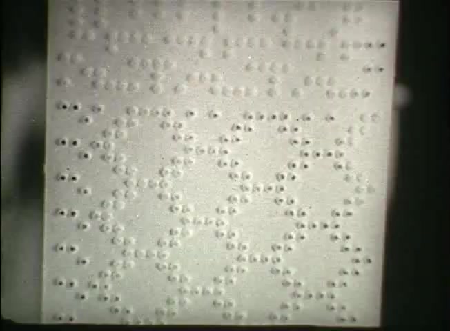 The still from 'Story Without End' features a close-up of a punched paper tape, showcasing a pattern of holes that represent data or commands. This visual emphasizes early telecommunication technology used by the Bell System in the late 1940s, illustrating the methods of information transmission at the time. The arrangement of holes reflects the systematic and organized approach of the network's operational procedures.