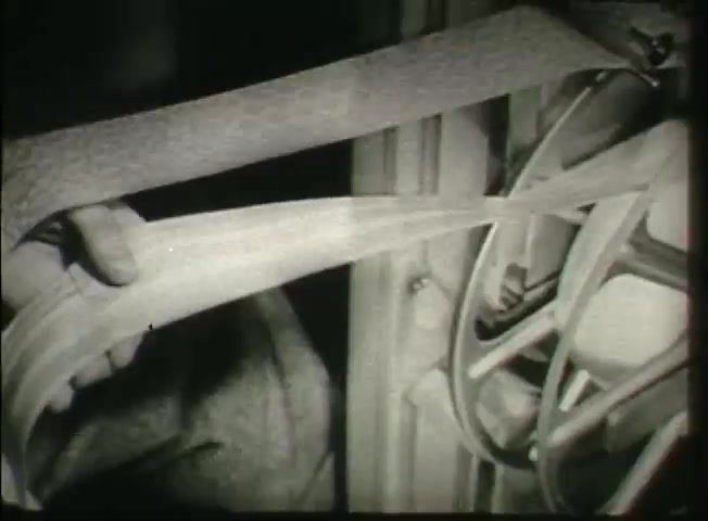 The still from 'Story Without End' captures a close-up view of a technician's hand manipulating a tape or ribbon within a central office equipment setup. The image showcases the intricate mechanics of telecommunications technology from the late 1940s, illustrating the era's focus on the operations of the Bell System. The monochromatic tones emphasize the industrial nature and complexity of the equipment, highlighting the behind-the-scenes work essential for network functionality.