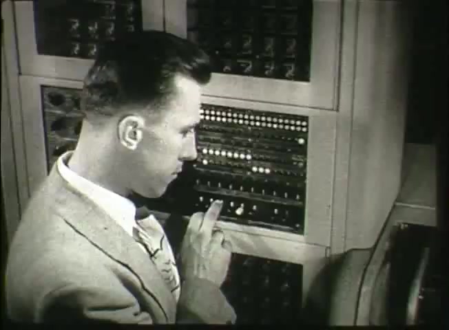 The still from 'Story Without End' features a man in a light-colored suit, closely examining and interacting with a telephone switching equipment panel. His focus suggests a technical role, possibly an operator or technician. The panel showcases a series of buttons and lights, indicative of mid-20th century telephone technology. This scene likely highlights the inner workings of the Bell System's network during the late 1940s, reflecting advancements in communication technology at the time.