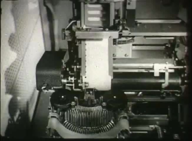 The still captures a close-up view of retro telecommunications equipment, likely used in a central office. The image showcases a mechanical device with moving parts, including a typewriter-like structure and rotating elements. It reflects the technological capabilities of the late 1940s, illustrating the operation of the Bell System's network during that era, with an emphasis on machinery involved in telephony processes. The monochrome aesthetic is characteristic of the period's film quality.