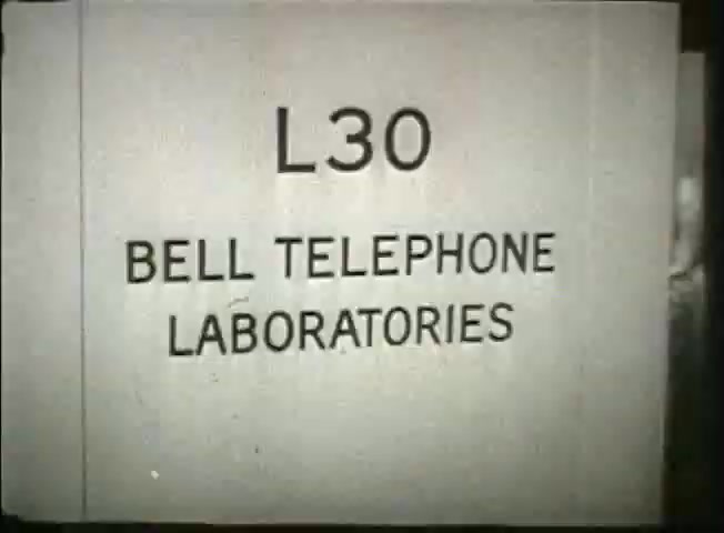 The still features a sign reading 'L30' and 'BELL TELEPHONE LABORATORIES,' indicating a specific location within the Bell System. This film segment from the late 1940s provides insight into the telecommunications network of the time, showcasing some central office operations and highlighting the technological advancements of the Bell Laboratories. The visual style reflects the era's documentary aesthetic.
