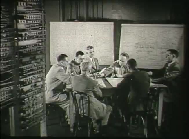 The still from 'Story Without End' features a group of six men gathered around a table in a central office setting, likely discussing telecommunications matters. They are dressed in business attire, indicative of the late 1940s. Behind them, a panel of electrical equipment is visible, highlighting the technological context of the Bell System at that time. The men appear engaged and focused, suggesting a collaborative effort to address network challenges. Whiteboards behind them contain notes and diagrams, adding to the technical atmosphere of the scene.