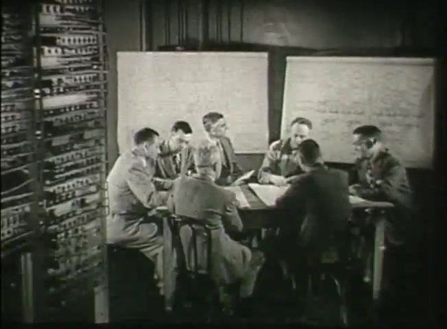 The still from 'Story Without End' features a group of six individuals gathered around a table in a central office setting, discussing or planning, with papers and charts in front of them. Behind them is a prominent switchboard, indicative of telephone technology from the late 1940s. The environment suggests a collaborative effort related to telecommunications, possibly outlining operational plans or innovations within the Bell System network during that era. Whiteboards with handwritten notes flank the group, further emphasizing the analytical or technical focus of their meeting.