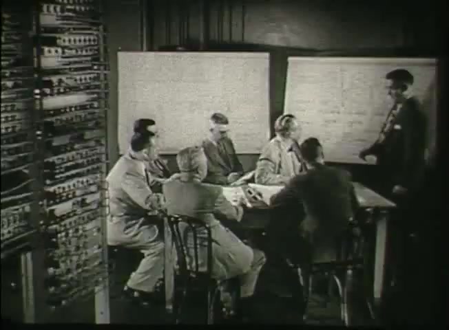 The still from 'Story Without End' features a group of seven men seated around a table in a central office environment. They appear to be engaged in a discussion, with one individual standing and presenting information, possibly related to the Bell System's network. Behind them, detailed equipment, likely telephone switching apparatus, is visible on the left. The setting reflects a mid-20th-century technological workspace, indicative of the era's focus on communication infrastructure.