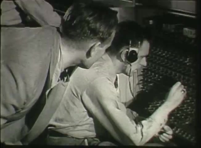 The still from 'Story Without End' features two men in a central office setting, typical of the late 1940s Bell System context. One man is wearing a headset and attentively operating a switchboard, while the other leans in to observe. This scene captures the process of telephone connections during that era, highlighting the human element in telecommunications before automation. The overall tone reflects the importance of the workforce in maintaining network operations.