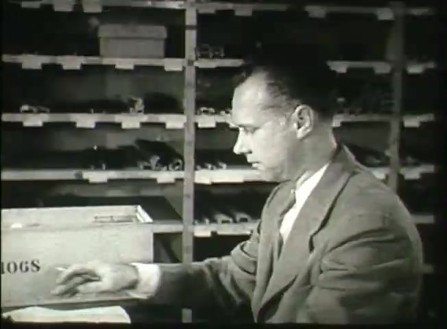 The still from 'Story Without End' depicts a man in a suit seated at a desk, focused on his work. He is writing or handling documents, with a box labeled '106S' visible in the foreground. Behind him is a shelving unit filled with various items, hinting at an organized workspace typical of a central office environment. The overall tone reflects the mid-20th century style, aligning with the film's purpose of providing insights into the Bell System network during that era.