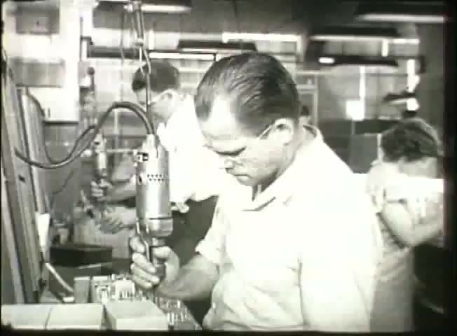 The still from 'Story Without End' shows a working environment likely in a central office during the late 1940s. In the foreground, a male technician is focused on using a handheld tool, possibly for assembly or repair. The background features additional workers engaged in similar activities, highlighting the collaborative nature of telecommunications operations at the time. The scene captures the essence of the Bell System's technological and workforce dynamics during that era.