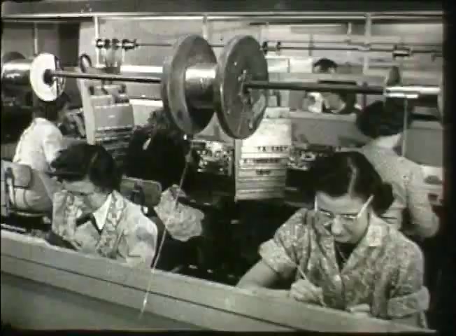 The still from 'Story Without End' features a bustling central office environment from the late 1940s, showcasing female workers engaged in telephone equipment assembly or maintenance tasks. Prominent in the foreground are two women focused on their work, with one using a pen and the other appearing to use tools. The background is filled with machinery and equipment typical of the Bell System, highlighting the era's technological landscape and the role of women in the telecommunications industry.