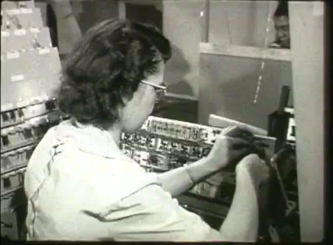 The still from 'Story Without End' features a woman working in a central office environment from the late 1940s, likely focusing on telephone operations or network systems. She is wearing glasses and engaged with a series of electrical circuits or connections, which reflects the technology of the time. In the background, another individual is partially visible, suggesting a collaborative work setting. The image captures a moment of hands-on technical work, highlighting the role of women in telecommunications during that era.