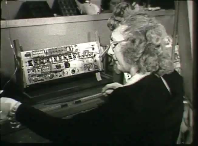 The still from the Bell System film 'Story Without End' features a woman, likely a telephone operator, engaged with a vintage switching console. The console shows a complex arrangement of circuits and wires, typical of the late 1940s technology. The operator, wearing glasses and with curly hair, appears focused as she interacts with the equipment, illustrating the human element of telephone operations during that era. This image captures a moment in the evolution of telecommunications, reflecting the network's functionality and the workforce behind it.
