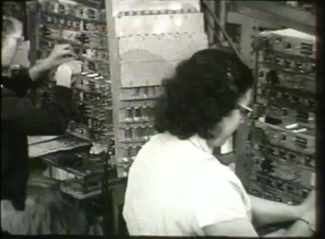 The still from *Story Without End* depicts two operators working in a central office environment, typical of the late 1940s Bell System. It shows a woman seated, engaged with telephone switchboards, while another operator in the background is adjusting equipment. The scene highlights the manual intricacies of telecommunications at the time, illustrating the human effort involved in managing calls within the network. The film aims to provide insight into the operational processes and significance of the Bell System during that era.