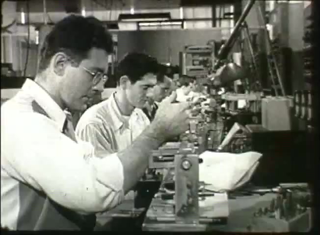 The still from 'Story Without End' depicts a factory-like setting where several men are engaged in meticulous work at assembly stations. The foreground features two workers, one with glasses, focused on their tasks, which likely involve intricate machinery related to telecommunications. The background hints at a bustling industrial environment, supportive of the Bell System's operations in the late 1940s. This captures a moment in the era's technical advancements and the dedication of its workforce.