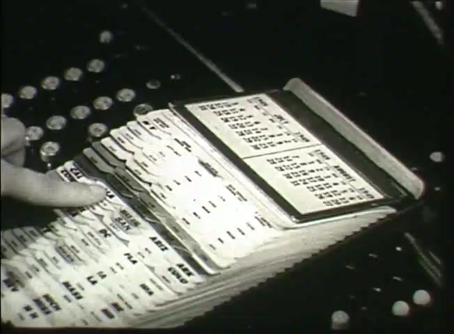 The still from 'Story Without End' features a close-up of a telephone switchboard interface, showcasing a stack of cards with printed information. A hand is visible, indicating interaction with the cards, likely related to call routing or network management. The background includes buttons or switches, typical of mid-20th-century central office equipment, reflecting the operational aspects of the Bell System during that era. This scene highlights the manual processes used in telecommunications before the advent of modern automation.