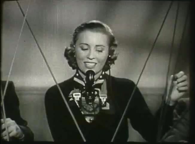 The still from *Story Without End* features a woman speaking into a vintage telephone microphone, depicting the communication technology of the late 1940s. She is dressed in a stylish outfit typical of the era, radiating a cheerful demeanor. The image suggests a focus on telecommunication, likely emphasizing the human element in the Bell System's network, possibly highlighting its role in connecting people. The presence of visible phone lines adds to the depiction of communication infrastructure at the time.