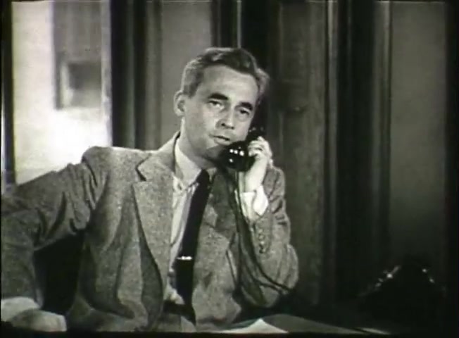 The still from 'Story Without End' features a man dressed in a light-colored suit and tie, sitting at a desk while holding a telephone to his ear. He appears engaged in conversation, looking directly ahead with a thoughtful expression. The background suggests an office setting, likely indicative of the late 1940s. This scene contributes to the film's exploration of the Bell System, highlighting personal communication and the infrastructure of the telephone network during that era.