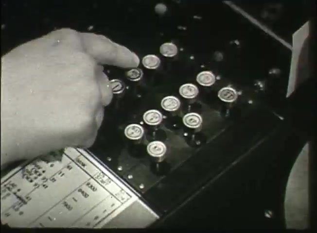 The still from 'Story Without End' features a close-up of a hand pressing a button on a control panel with several small, circular switches or buttons. The panel appears to be part of a vintage telephone central office system, indicative of the late 1940s technology used by the Bell System. Beneath the buttons, a printed layout or schematics is visible, likely containing operational instructions or network information relevant to the telephone system at that time. This snapshot encapsulates the historical context of telecommunication advancements during that era.