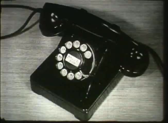 The still captures a classic black rotary telephone, a notable communication device from the late 1940s, prominently displayed on a surface. The phone features a circular dial with numbered and lettered segments, characteristic of the era before touch-tone technology. This image likely reflects the period's telecommunications infrastructure, illustrating the simplicity and functionality of the equipment used by the Bell System during that time.