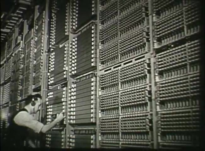 The still from 'Story Without End' depicts a technician working in a telephone central office from the late 1940s. The focus is on large, complex switching equipment, showcasing the intricate machinery of the Bell System's network at the time. The technician, wearing headgear, appears to be engaged in maintenance or operation, highlighting the human element in managing telecommunications infrastructure. The scene reflects the technological advancements and the importance of communication systems in that era.