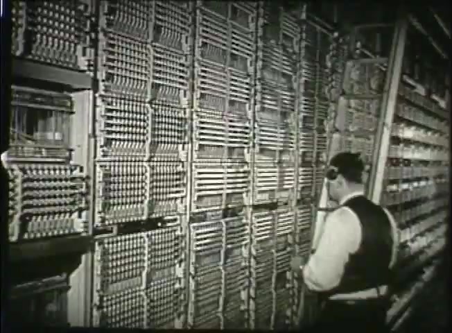 The still from 'Story Without End' captures a technician in a central office of the Bell System, standing in front of a large switchboard. The intricate network of wires and connections reflects the telecommunications technology of the late 1940s. The technician, wearing a headset, appears focused on monitoring or adjusting the equipment, highlighting the complexity and crucial nature of phone network operations during that era.
