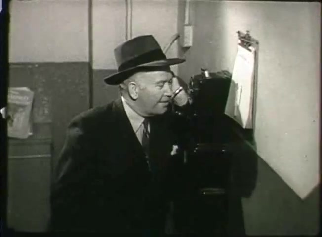 The still from 'Story Without End' features a man in a suit and hat speaking on a vintage telephone. The setting appears to be a central office with a plain wall and a clipboard visible nearby. This scene is part of a Bell System film from the late 1940s, likely illustrating the operational aspects of the telephone network during that era. The overall atmosphere conveys a sense of professionalism and the importance of communication technology at the time.