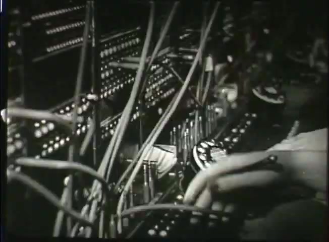 The still from 'Story Without End' presents a close-up view of a telephone switching board typical of the late 1940s. It features an array of illuminated buttons and complex wiring, showcasing the manual operation of the Bell System's central office. A hand is seen interacting with the equipment, illustrating the labor involved in traditional telecommunication systems. This snapshot captures the technological landscape and operational practices of the time, emphasizing the intricacies of network management before the advent of automated systems.