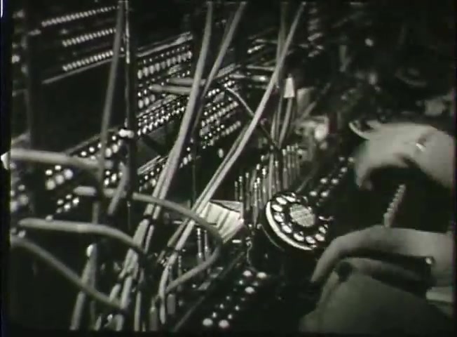 The still from the Bell System film 'Story Without End' depicts a close-up view of a telephone switching system from the late 1940s. It features a tangle of wires and numerous buttons, highlighting the complexity of the telecommunications network at that time. A hand is seen operating a rotary dial, emphasizing the manual nature of phone connections before the advent of modern technology. The black-and-white imagery adds to the historical context, reflecting the era's technological landscape.