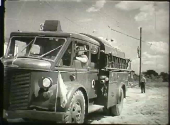 The still from 'Story Without End' showcases a vintage bus marked with a flag and a Bell System logo, likely indicating its use for communication purposes during the late 1940s. It features a driver at the wheel, with a rural backdrop that includes power lines and a person standing nearby. The image reflects the technological context of the time, highlighting the Bell System's role in network development.