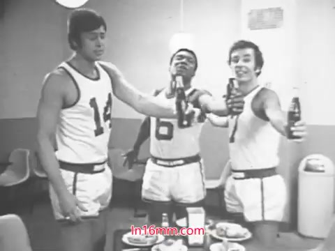 The still from the Diet Rite Cola commercial features three basketball players in casual sports attire, suggesting a relaxed, celebratory atmosphere. They are gathered around a table filled with food, raising bottles of Diet Rite Cola in a toast. The commercial likely emphasizes the refreshing qualities of the drink, especially in light of the formula change from cyclamate to saccharine around 1969. The presence of Hondo Havlicek, a notable player from the Boston Celtics, adds to the appeal, linking the product to professional basketball and an active lifestyle.