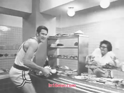 The still from the Diet Rite Cola commercial features John 'Hondo' Havlicek, a Boston Celtics player, in a playful scene. He is wearing a basketball uniform and holding a drink, likely Diet Rite Cola, while engaging with a character in a white robe behind a dessert display. The setting gives off a lighthearted, humorous vibe reflective of advertising from that era. The commercial aligns with Diet Rite's 1969 formula change, aiming to highlight the beverage's appeal amidst a backdrop of indulgent snacks.