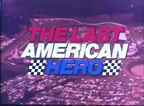 The still features the title 'THE LAST AMERICAN HERO' prominently displayed in bold, colorful letters—red, white, and blue—with checkered flag motifs flanking the text. The background shows an aerial view of a racetrack surrounded by a large crowd, indicative of a motorsport event. This composition sets the tone for a dramatic narrative focused on racing and heroism.