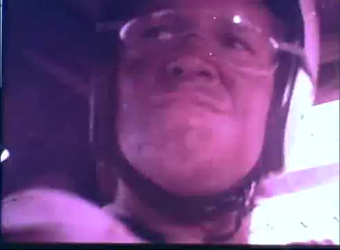 The still features a close-up of a determined male driver in a racing helmet and goggles. His expression conveys focus and intensity, reflecting a moment of adrenaline-fueled concentration. The color tone suggests a vintage aesthetic, typical of 16mm film, enhancing the nostalgic feel associated with the trailer for 'Last American Hero.' The background hints at a racing environment, with blurred details suggesting motion.