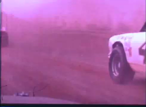 The still captures a dynamic racing scene from the 'Last American Hero' trailer, showing cars speeding through a dusty track. The focus is on the close-up of a white car, hinting at intense competition. The purple tint adds a nostalgic feel, typical of 16mm film aesthetics, enhancing the sense of motion and excitement inherent in the narrative. Dust swirls around, emphasizing the rugged and adrenaline-fueled environment of stock car racing.