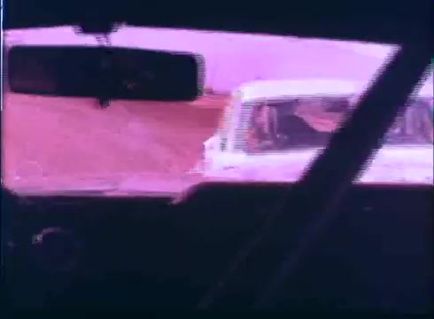 The still from the 'Last American Hero' trailer shows an interior view of a car, captured from the driver's perspective. The focus is on the rearview mirror, which reflects a white car behind. The image has a distinctive color tone, likely due to the 16mm film stock used, conveying a vintage aesthetic. The background hints at a dusty road, enhancing the movie's racing theme.