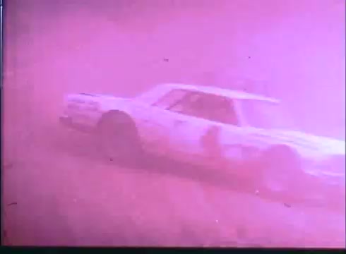 The still captures a race car in motion, surrounded by a smoky, hazy atmosphere, likely depicting a dramatic moment from the film's racing scenes. The car, likely white with a number on the side, appears to be navigating a challenging track, emphasizing the excitement and intensity of the racing theme in 'Last American Hero.' The pink tint adds to the gritty, stylized feel of the trailer.