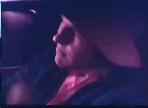 The still shows a scene from the trailer of 'Last American Hero.' A man with a serious expression is seated in a car, partially illuminated, suggesting a tense or reflective moment. The atmosphere is somewhat moody, enhanced by muted colors typical of 16mm film. This scene likely captures his character's inner conflict or determination.